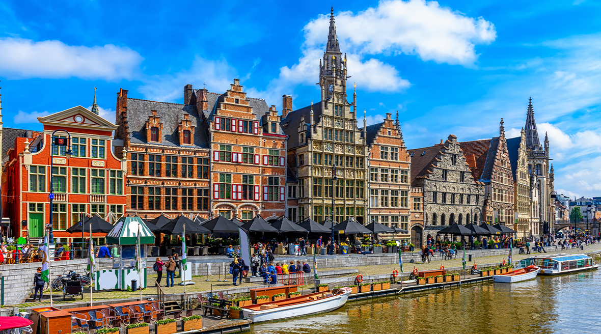 cityscape of Ghent with intricate buildings on the riverbank with people walking by and enjoying shops and restaurants